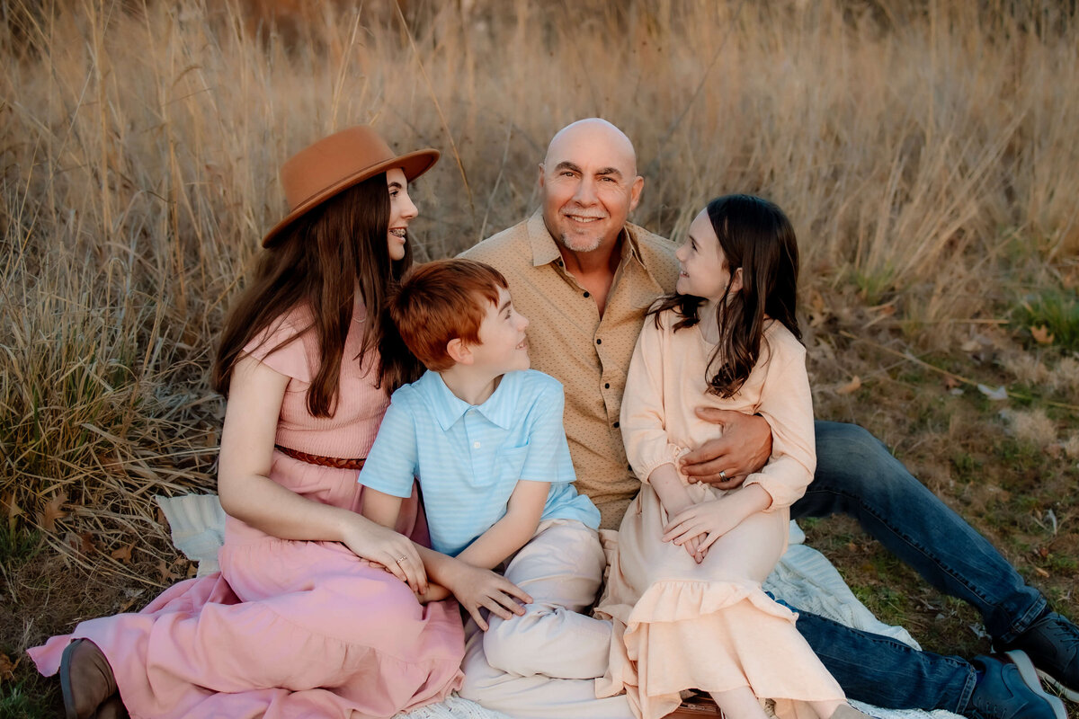 A father sitting on a blanket with his 2 daughters and son in a field at sunset