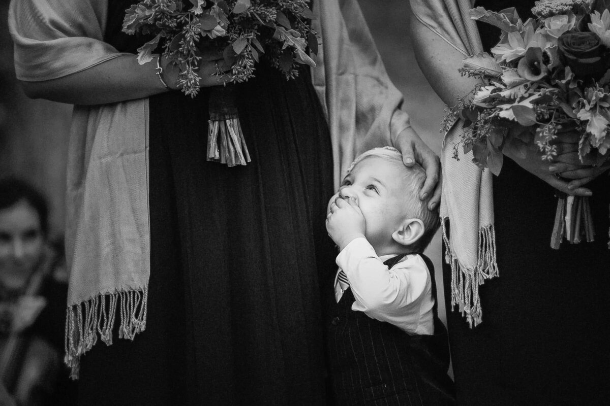 A young child peeks out curiously between two bridesmaids, with a whimsical expression on his face during a wedding ceremony.