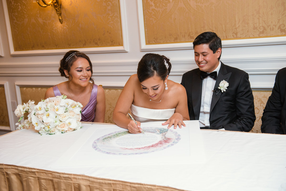 photo of bride signing the ketubah aside her groom and bridesmaid from wedding at The Garden City Hotel