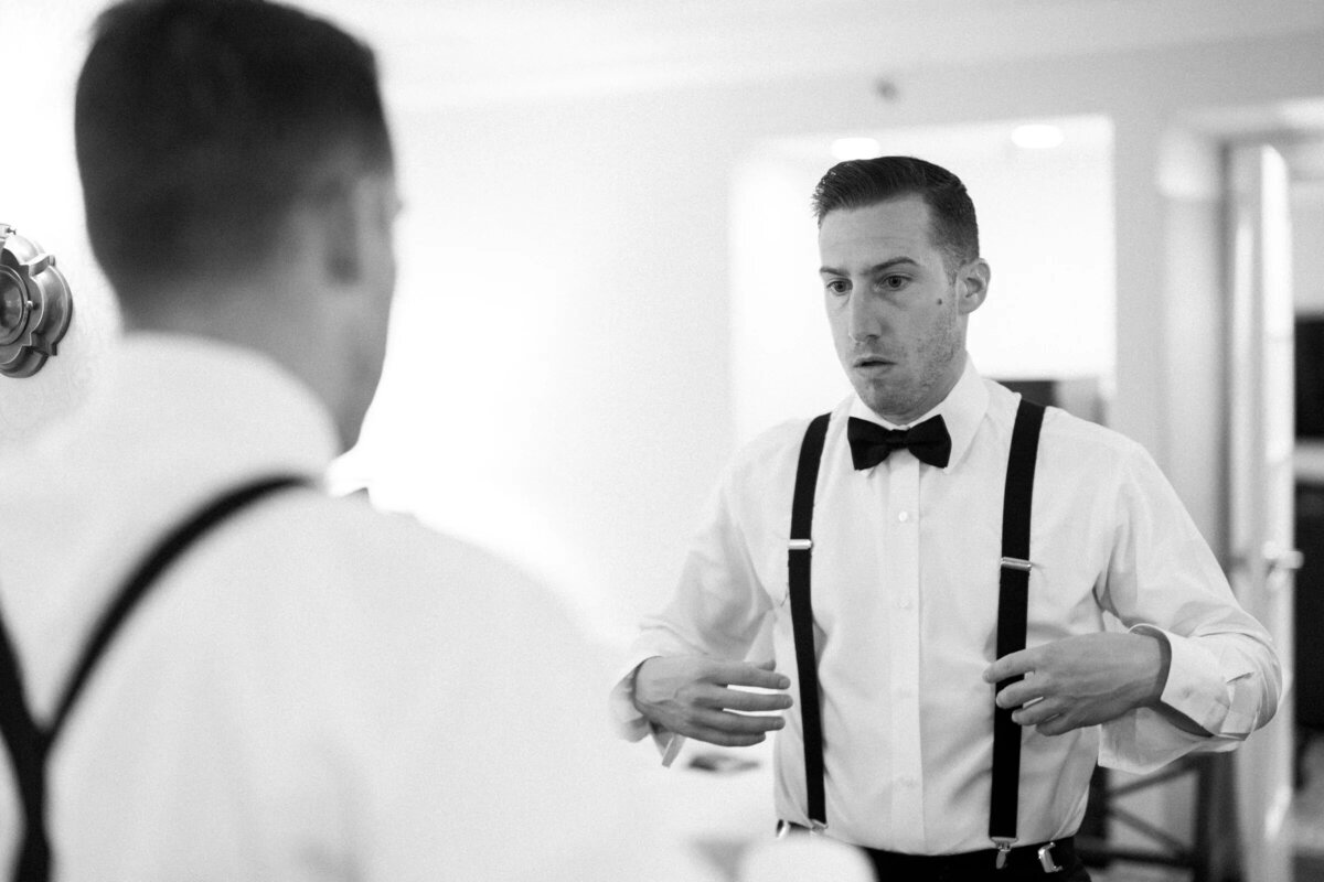 A black and white photo of a groom in suspenders making final adjustments in the mirror before the wedding ceremony.