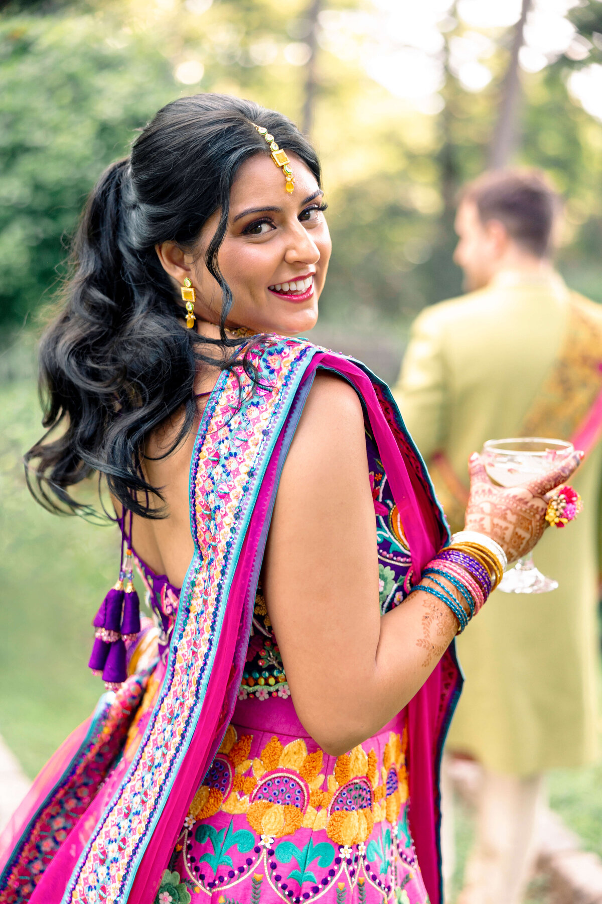 A woman in vibrant traditional attire smiles at the camera, holding a drink. Her outfit features intricate patterns and bright colors. She has henna on her hands and colorful bangles. A man in similar attire is blurred in the background.