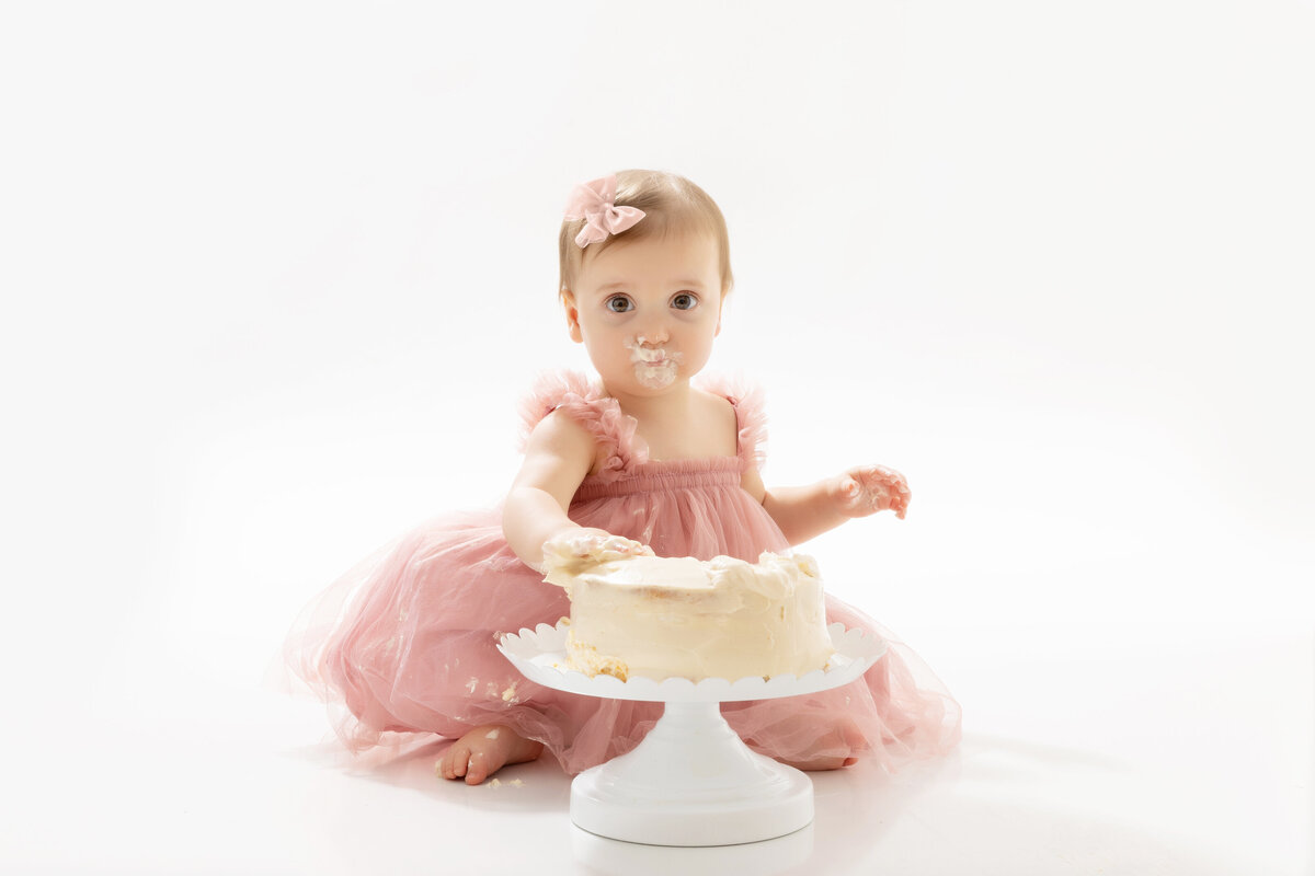 A toddler girl in a pink tutu sits in a white studio eating a cake