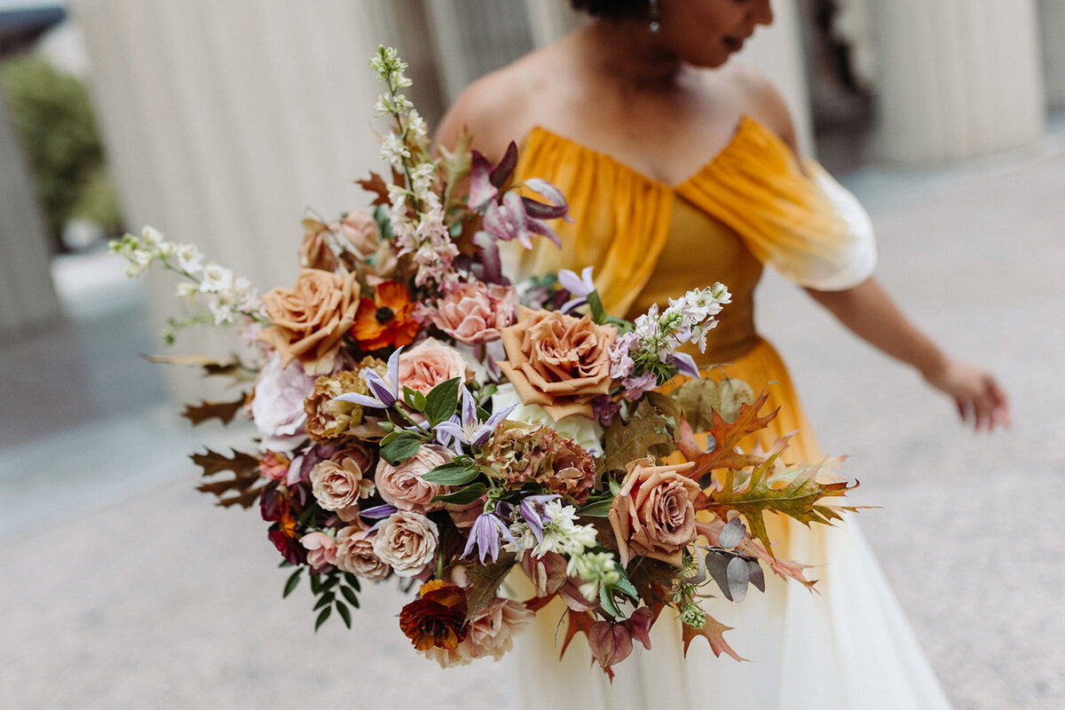 Lush bridal bouquet in autumnal hues of orange, burgundy, dusty rose, and lavender composed petal heavy roses, ranunculus, lavender clematis, and fall foliage. Design by Rosemary and Finch in Nashville, TN.