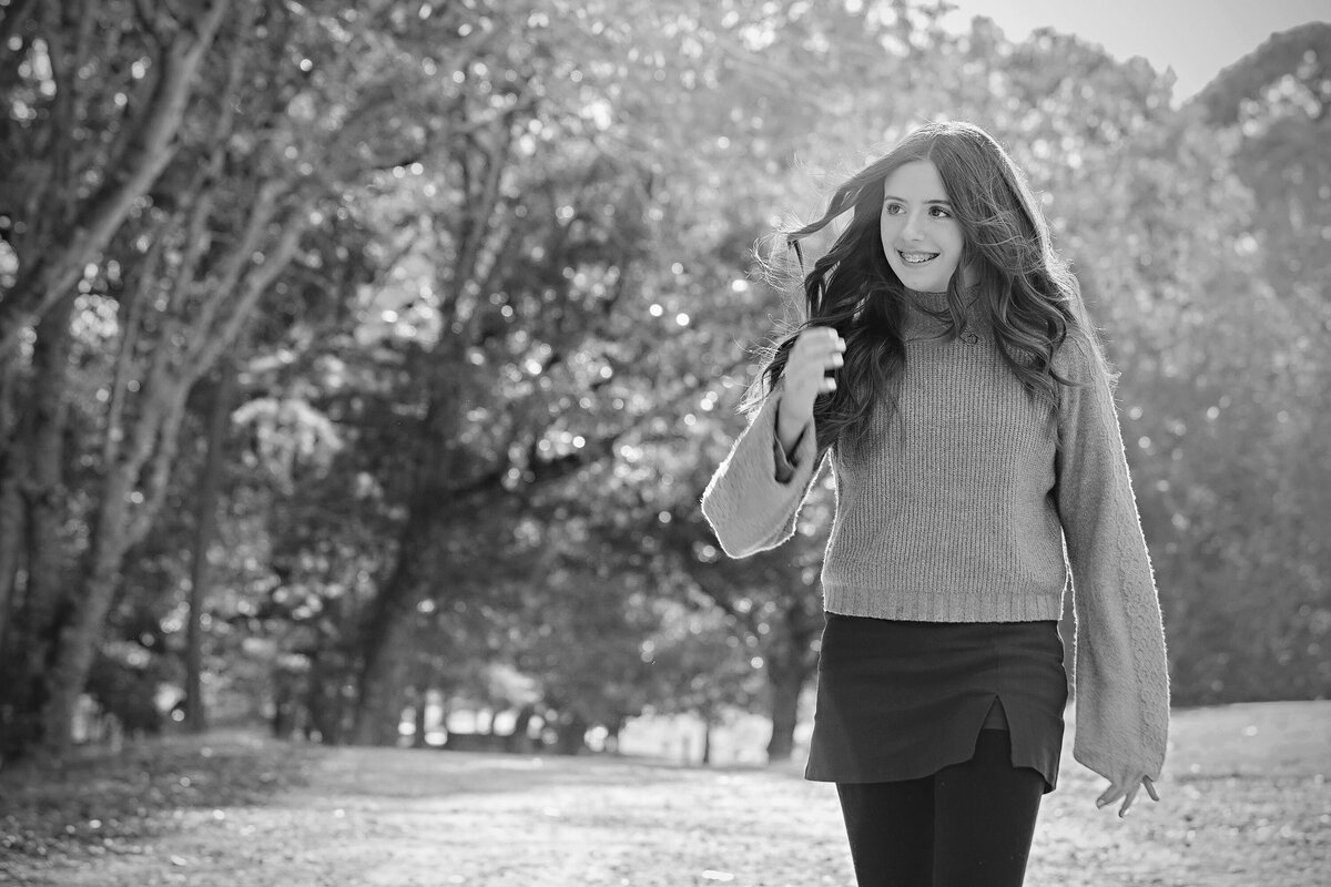 Sweet middle school girl in tan sweater and cranberry skirt standing on gravel roadway lined with fall foliage at Anne Springs Close Greenway Charlotte NC. Photo in black and white