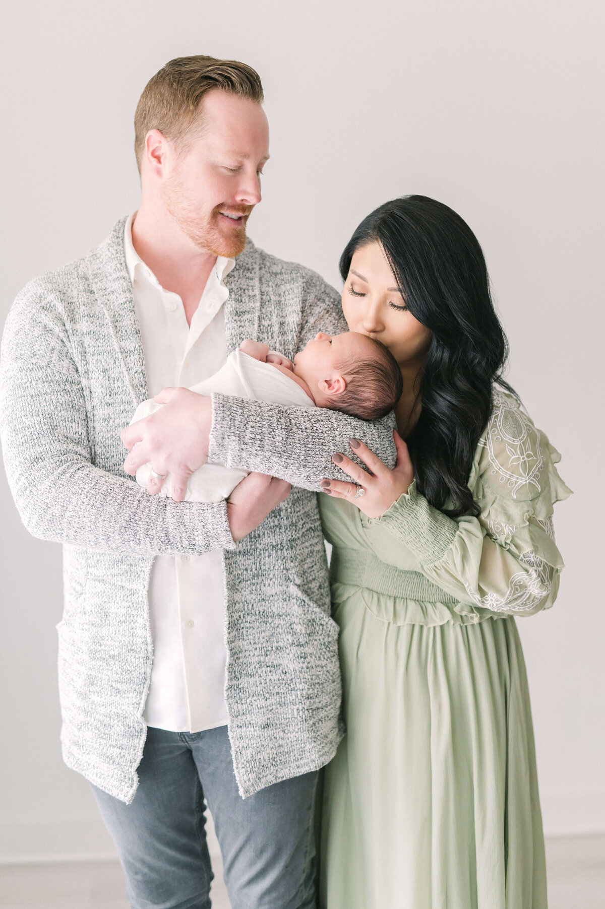 first time parents posing with newborn baby boy and mama kissing baby's head