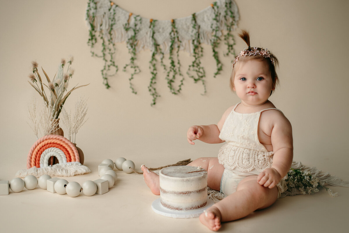 One year old girl with white iced birthday cake. She is wearing a white romper and sitting on cream backdrop with bohemian decorations behind her.