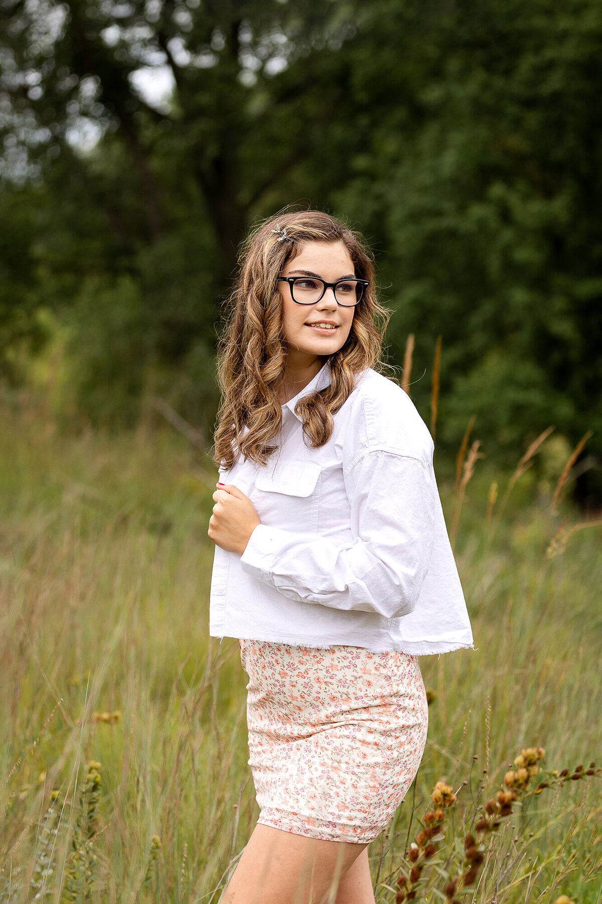 Beautiful girl with glasses wearing a floral dress with a white denim jacket walks among prairie grasses.