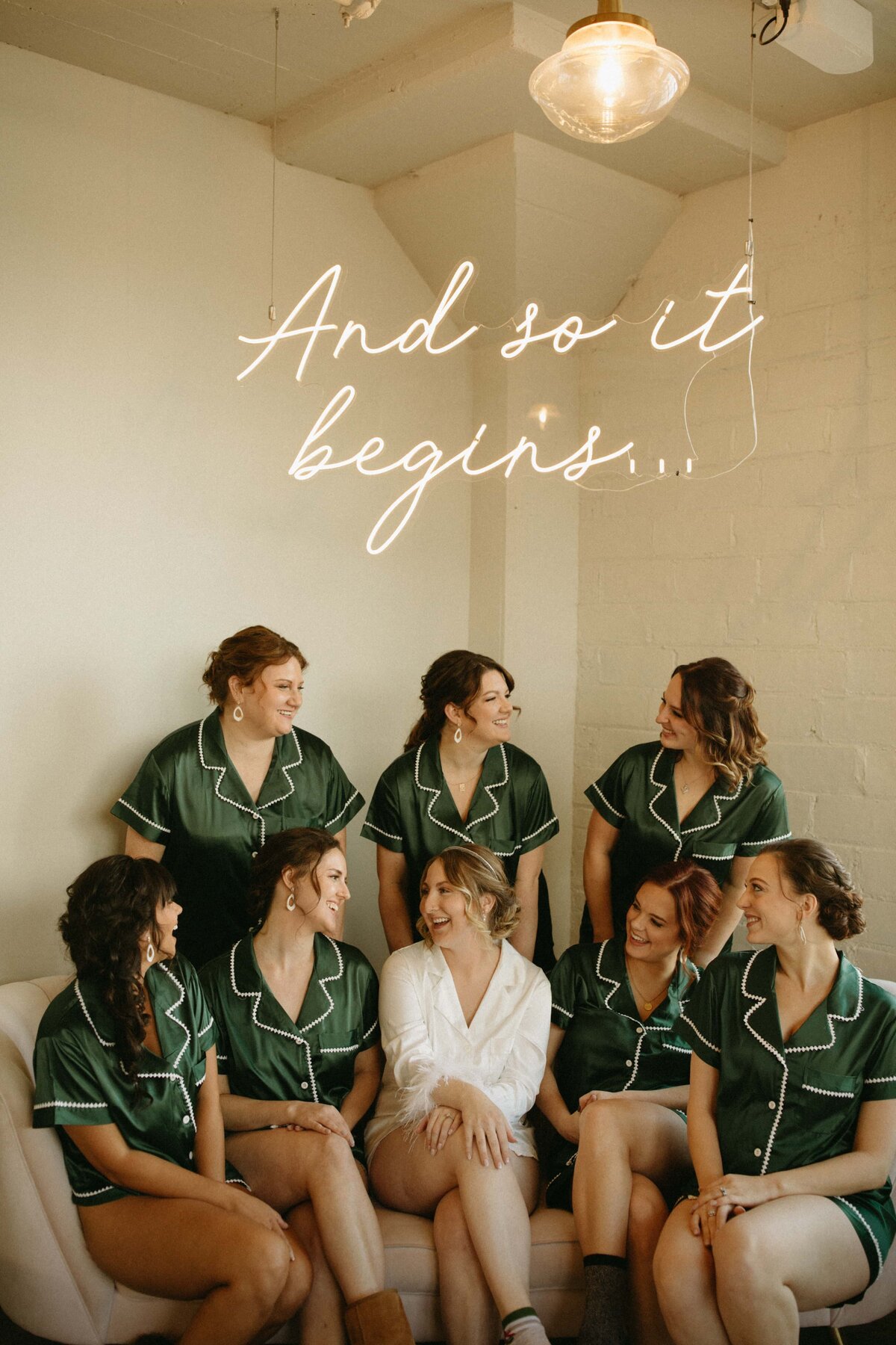 Bride wearing a white robe and bridesmaids in green robes smiling and sitting together under a neon sign at Park Farm Winery that reads "and so it begins...