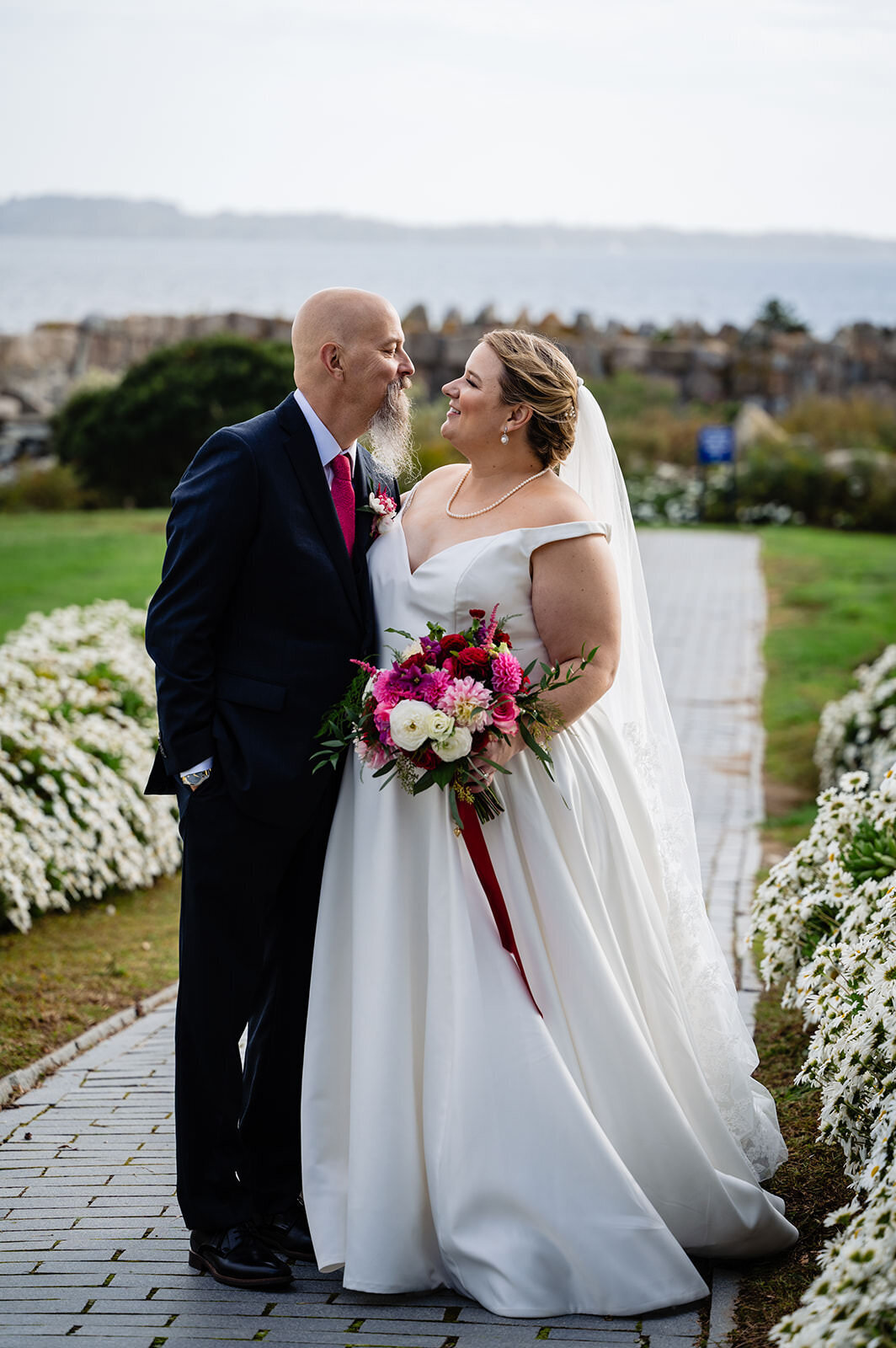 A bride and groom gaze into each other's eyes, standing amidst white flowers with a coastal backdrop