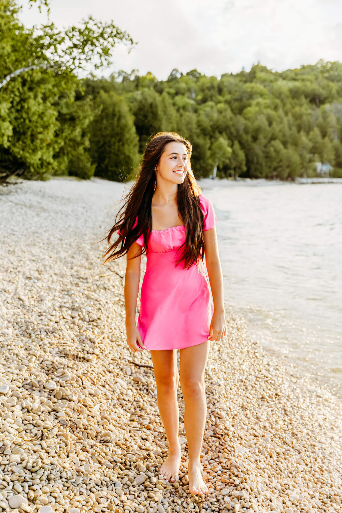 stunning young lady in a pink dress walking along the rocky shore in Door County for senior photography session