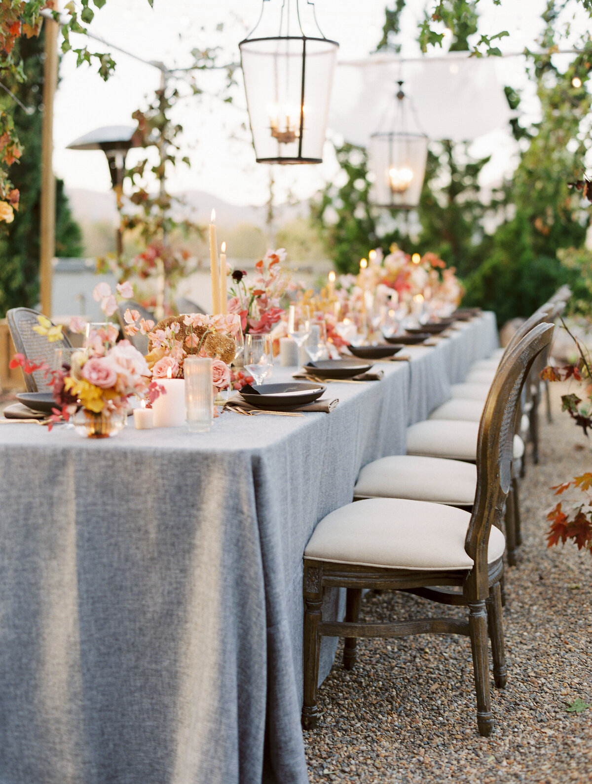 a wedding reception table with wooden chairs and light grey tablecloth with autumn colored florals and large lanterns