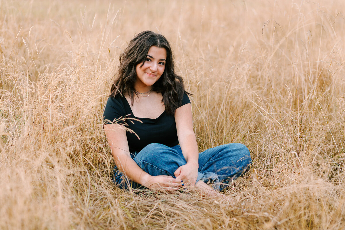 A teenage girl in a black t-shirt and jeans sits in a field smiling