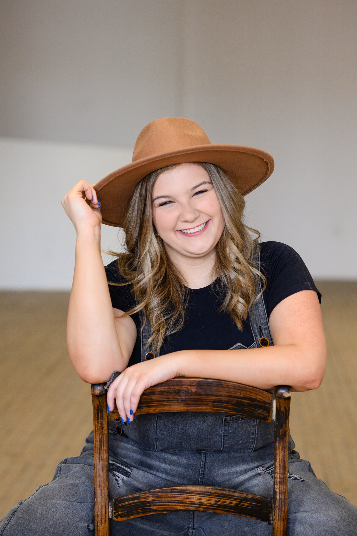 Senior photo outfit idea of a girl in black overalls sitting backward in a wooden chair with a brown hat
