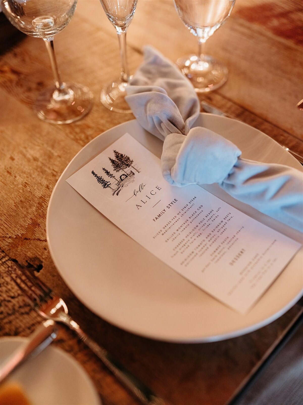 Candle-lit closeup of wedding reception tablescape, round white plate, elegant glasses and silverware, gray linen napkins and minimalist custom menu design.