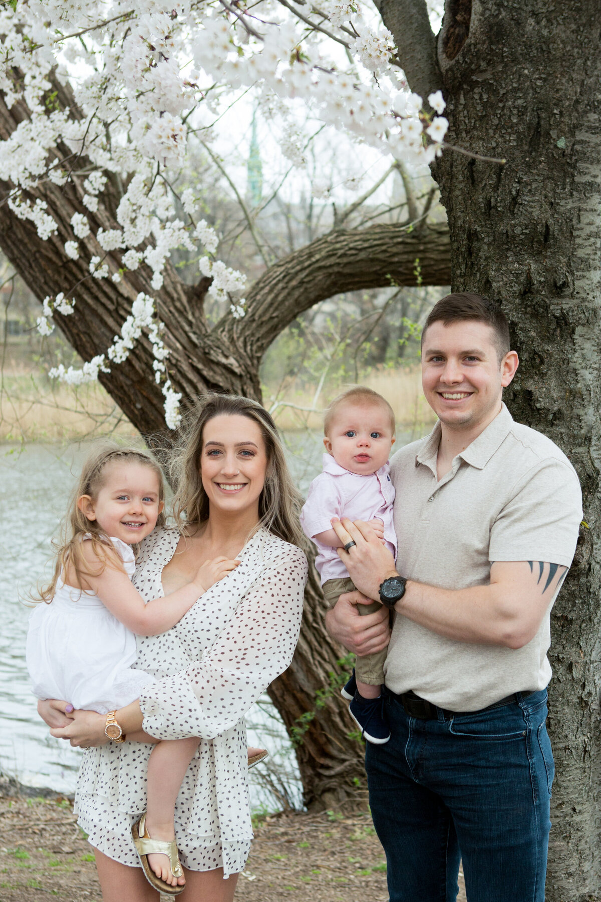 Family of four with a white mother in a polka dot romper and a dad in jeans and a khaki polo shirt hold their young children in front of a cherry blossom tree Branch Brook Park New Jersey mini-session photographer photographer Milestone Images