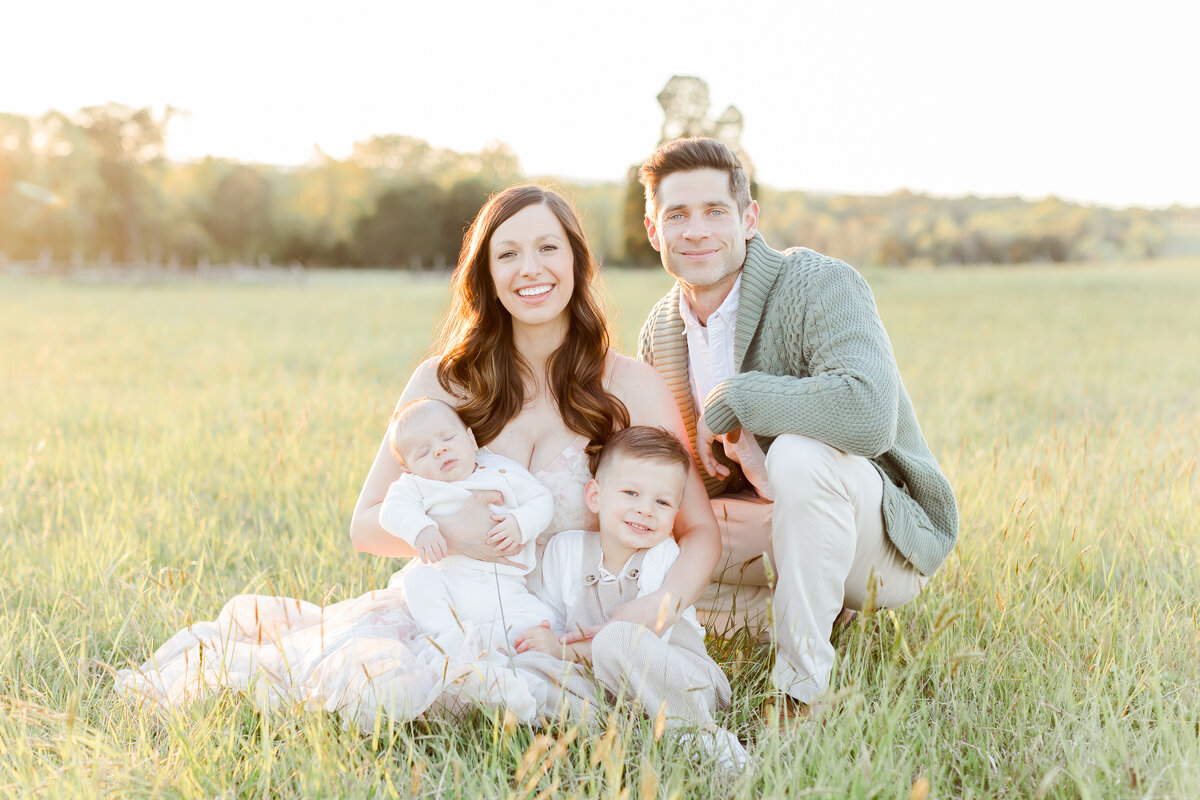 A family sitting down outside in a field smiling at the camera by Northern Virginia Family Photographer