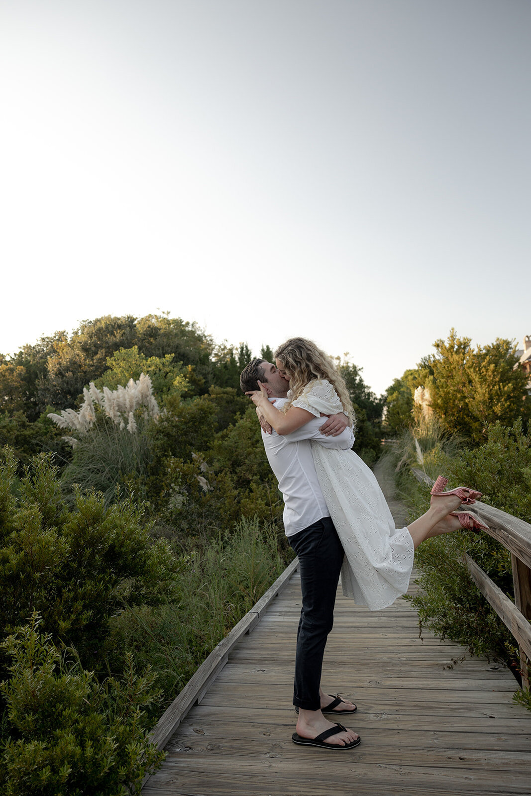 Man lifts up woman in white dress and shares a kiss. Both standing on beach boardwalk with greenery in background.
