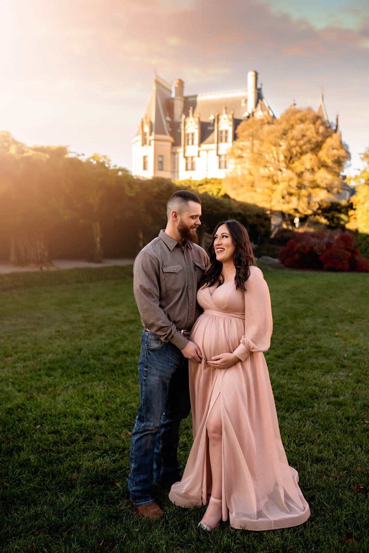 A beautiful mama to be in a long pink gown smiles at her husband while standing in front of the Biltmore Estate