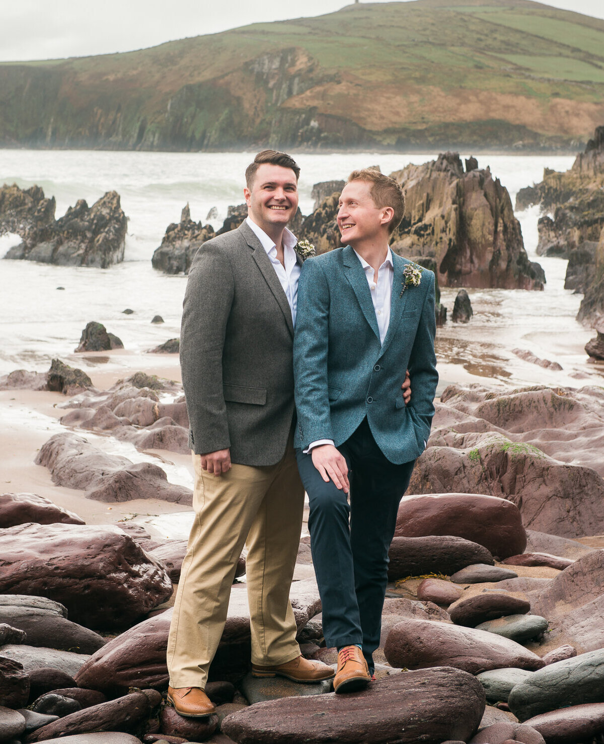 Two grooms standing on rocks looking at each other on beach