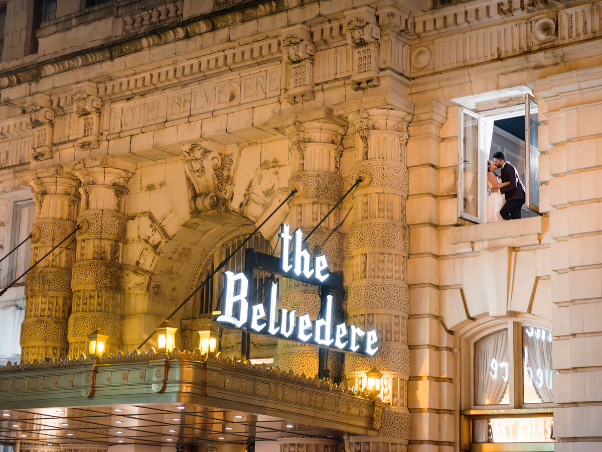 Historic building facade with ornate architecture, featuring a sign reading the Belvedere. Two workers are installing or repairing a window on the upper floor. The scene is illuminated by warm lights, creating a lively evening atmosphere.