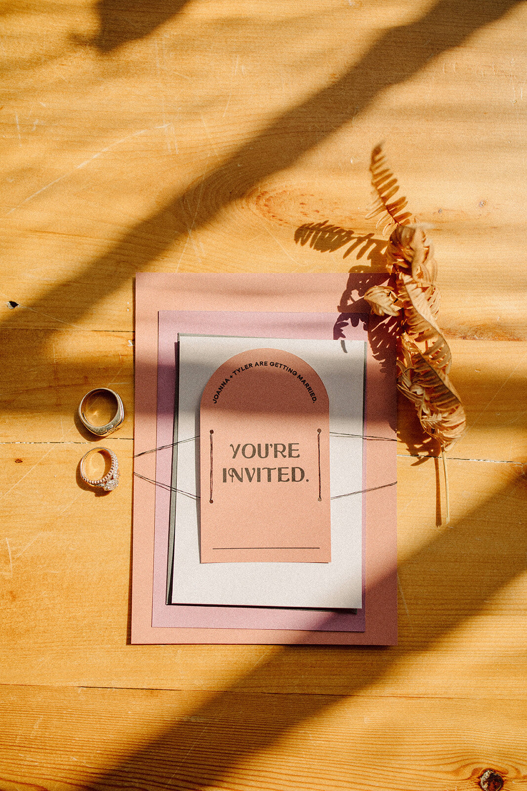 Rounded peach wedding invitation set atop off-white and mauve-colored wedding stationery and wedding bands atop wooden table.