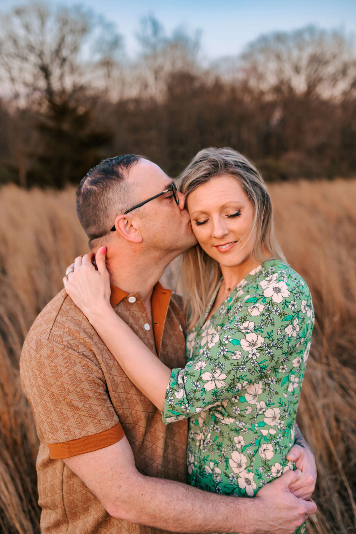 a man is kissing the temple of his wife, she is smiling and eyes closed , they are standing in a field of tall grass