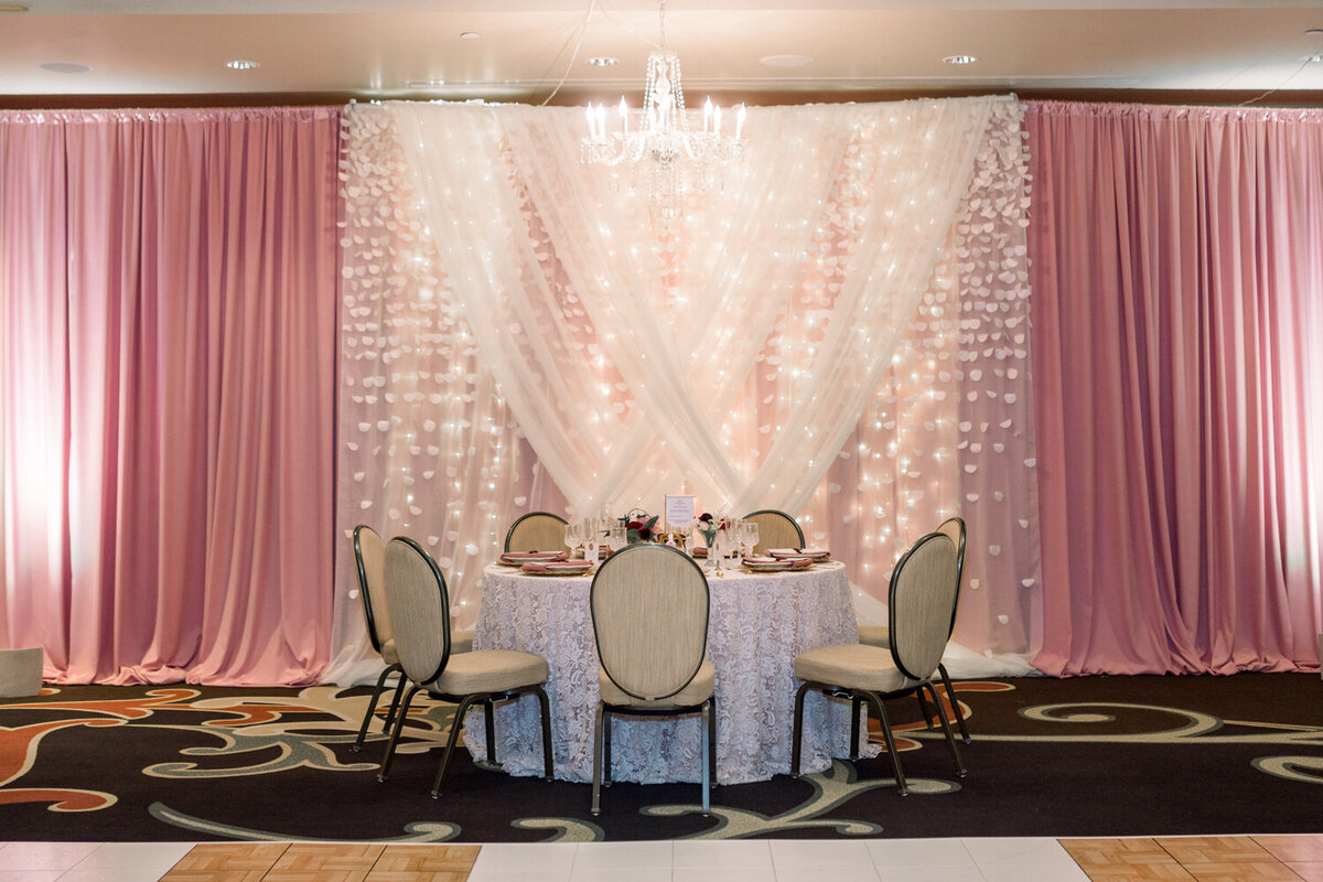 Head table at a 50th wedding anniversary celebration with dusty rose and sheer white drapes layered in a criss-cross design, enhanced by uplighting. The table features a white lace overlay and is adorned with burgundy and blush floral centerpieces, creating a refined and romantic ambiance at The Northern Hotel in Billings, MT.