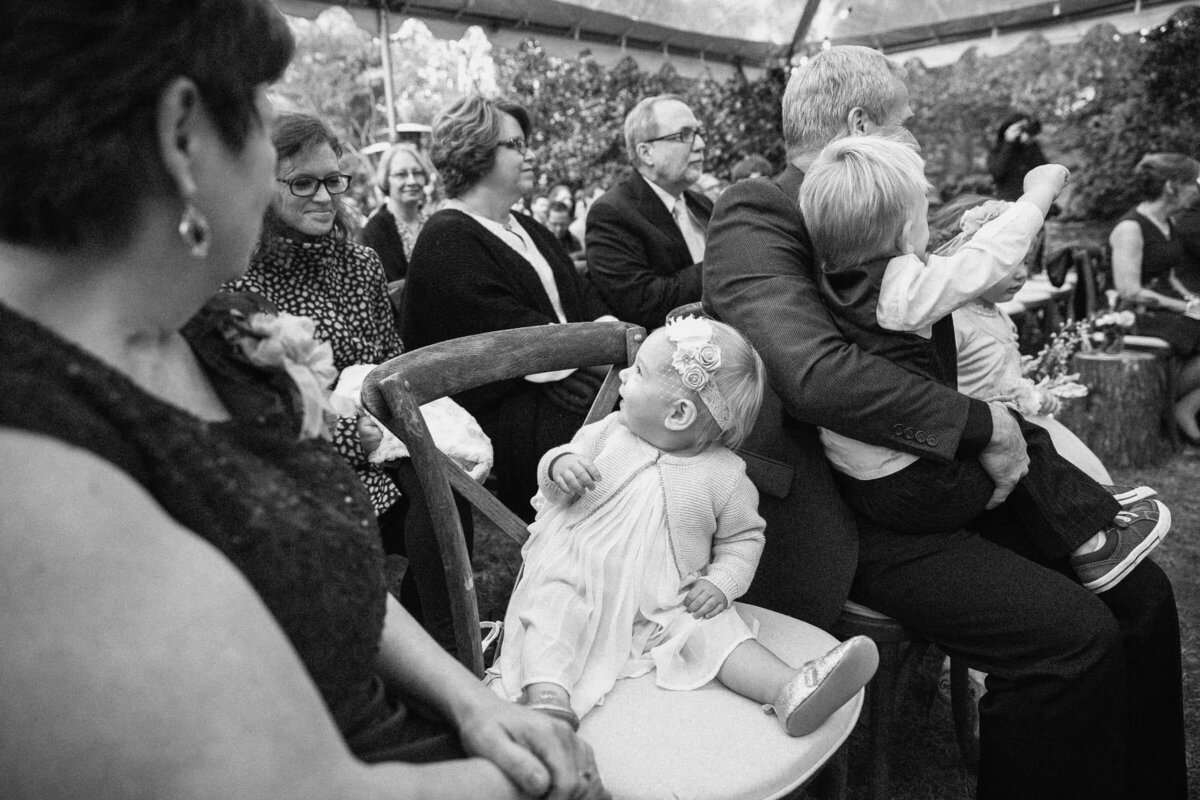 A black and white image capturing a cheerful baby looking up in awe, held by a guest at a wedding ceremony.