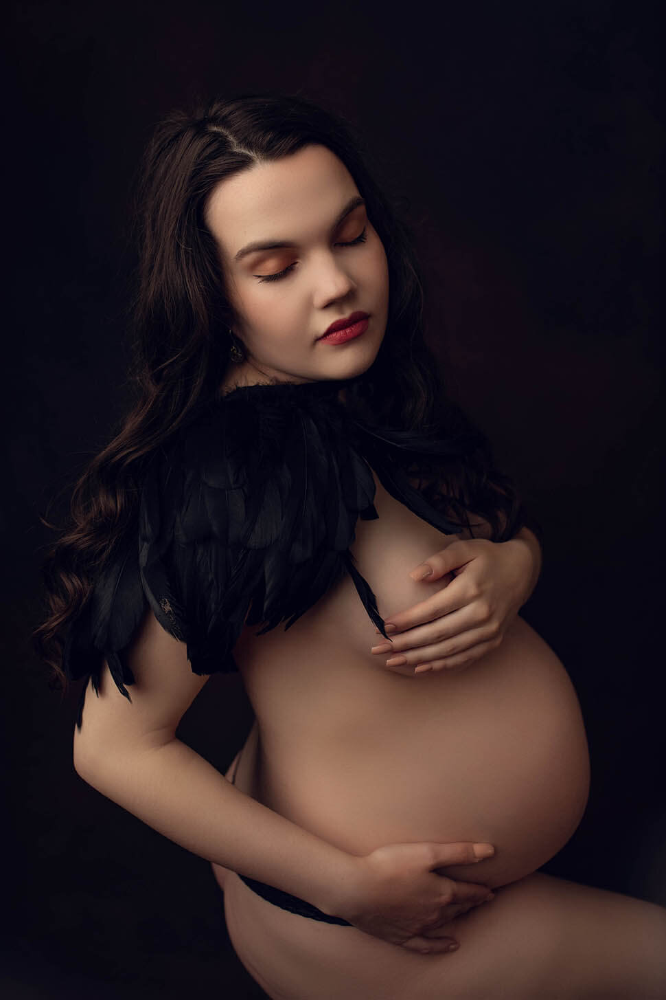 a nude woman holds her bump and breast while wearing a feather capelet and her eyes are closed