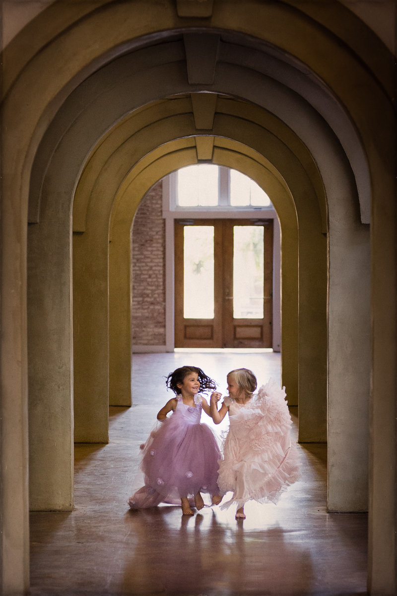 Two little girls wearing fancy gowns in pink and purple hold hands, laugh, and smile while running down hallway of Sky 9 Studio.