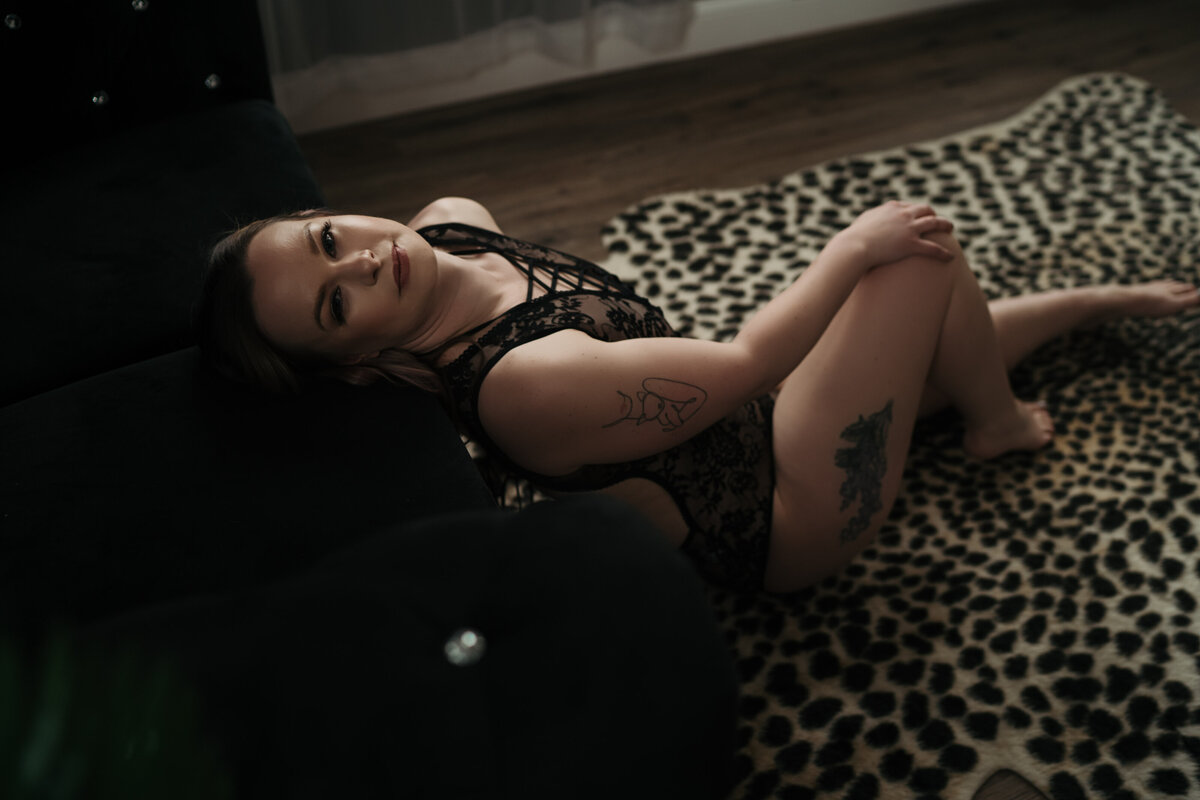 A woman in black lace lingerie leans back onto a black couch with a hand on her knee on a cheetah print rug