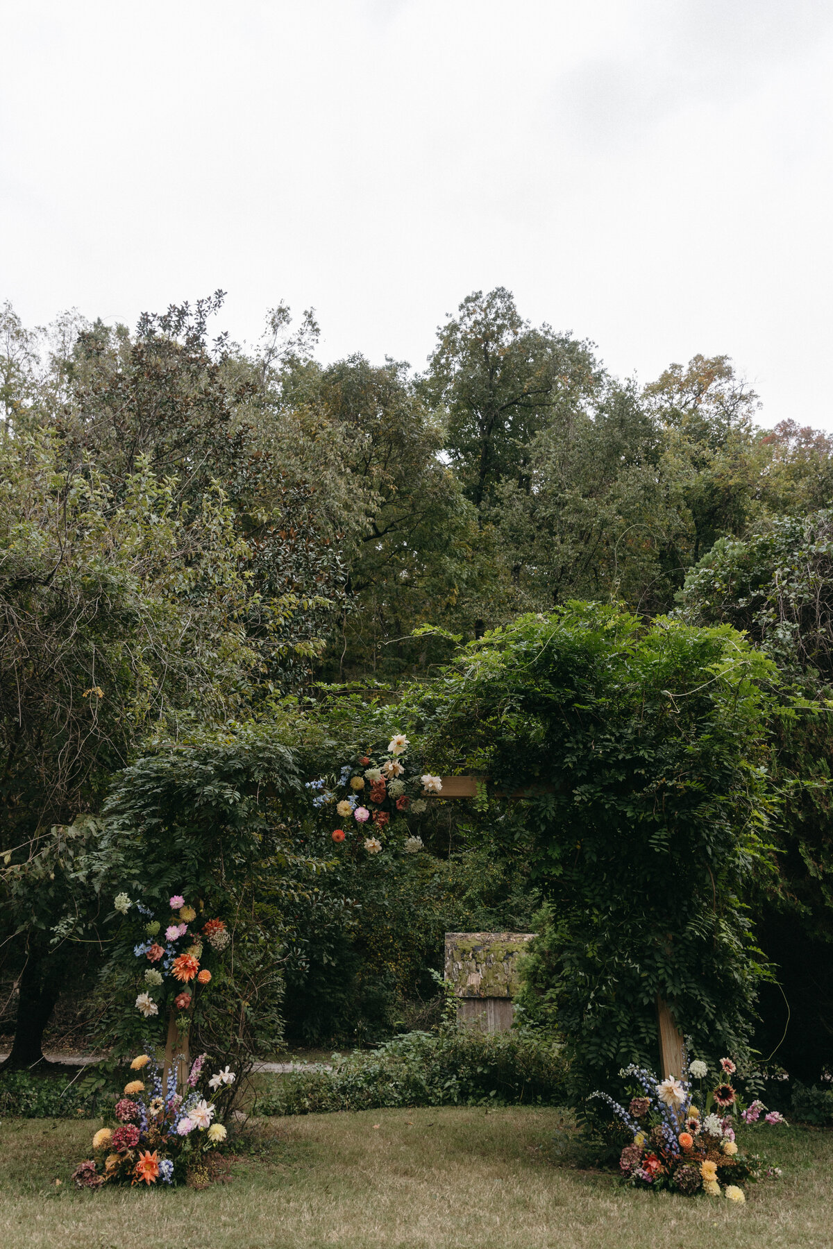 Garden wedding ceremony arch covered in lush greenery and vibrant florals including dahlias, roses, delphinium, and seasonal blooms for a whimsical natural backdrop.