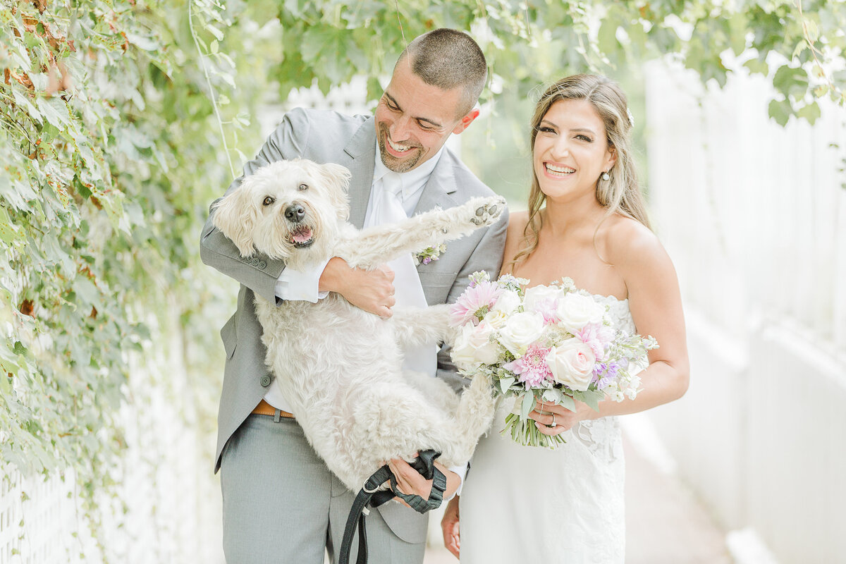 Bride and groom pose for a casual wedding portrait with their pup at the Five Bridge Inn in Rehoboth, MA. The groom is holding the dog and smiling at him. The bride is holding her bouquet and smiling at the camera. Captured by MA wedding photographer Lia Rose Weddings