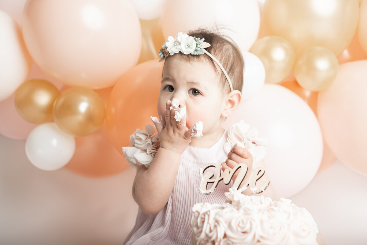 Birthday baby girl eating a messy pink cake with a pink peach white and gold balloon garland