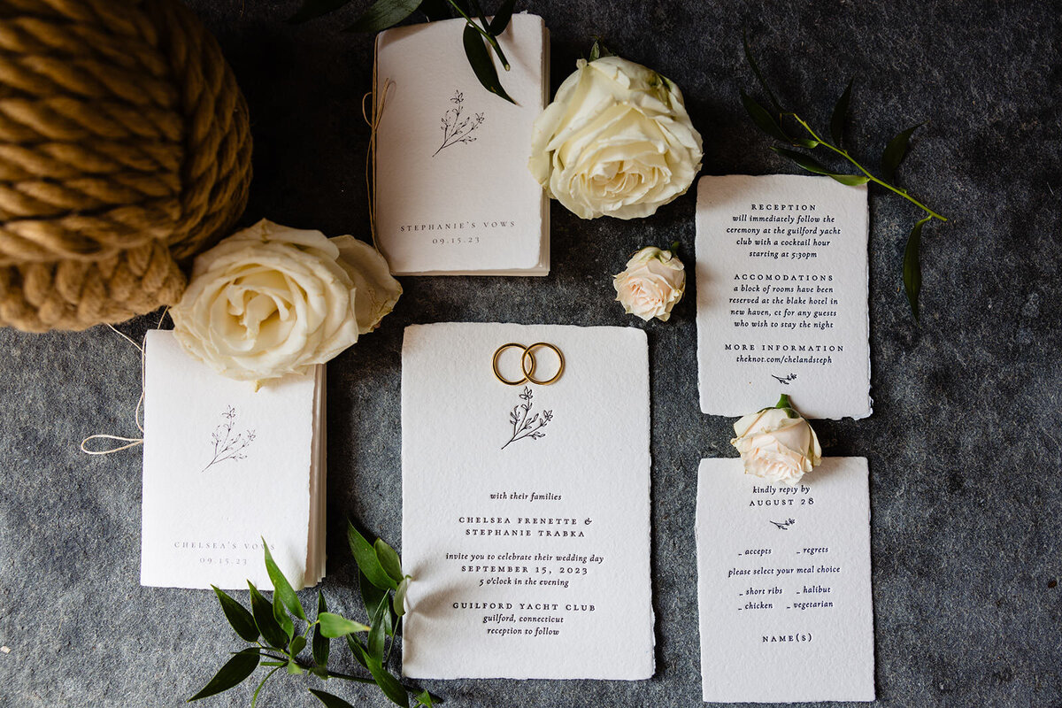 A wedding invitation suite laid out with rings, featuring elegant white and gold text on textured paper, accompanied by fresh roses and green leaves.