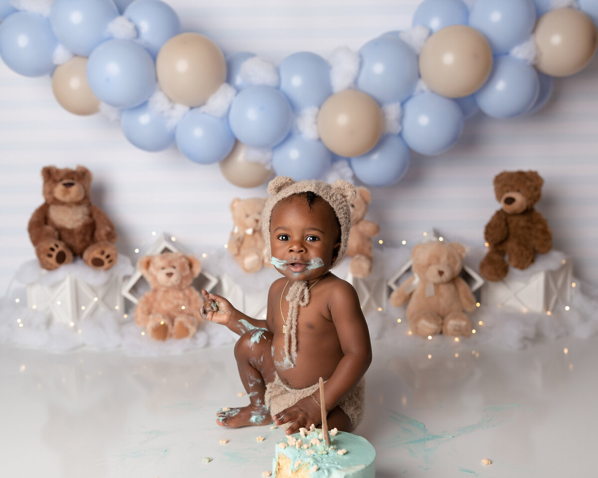 Teddy bear themed cake smash at West Palm Beach photography studio. Baby boy is wearing knit shorts and a matching teddy bear bonnet. He has cake on his face, hands, and legs and is looking at the camera. Behind him there is a blue and beige balloon arch with teddy bears sitting on white crates.