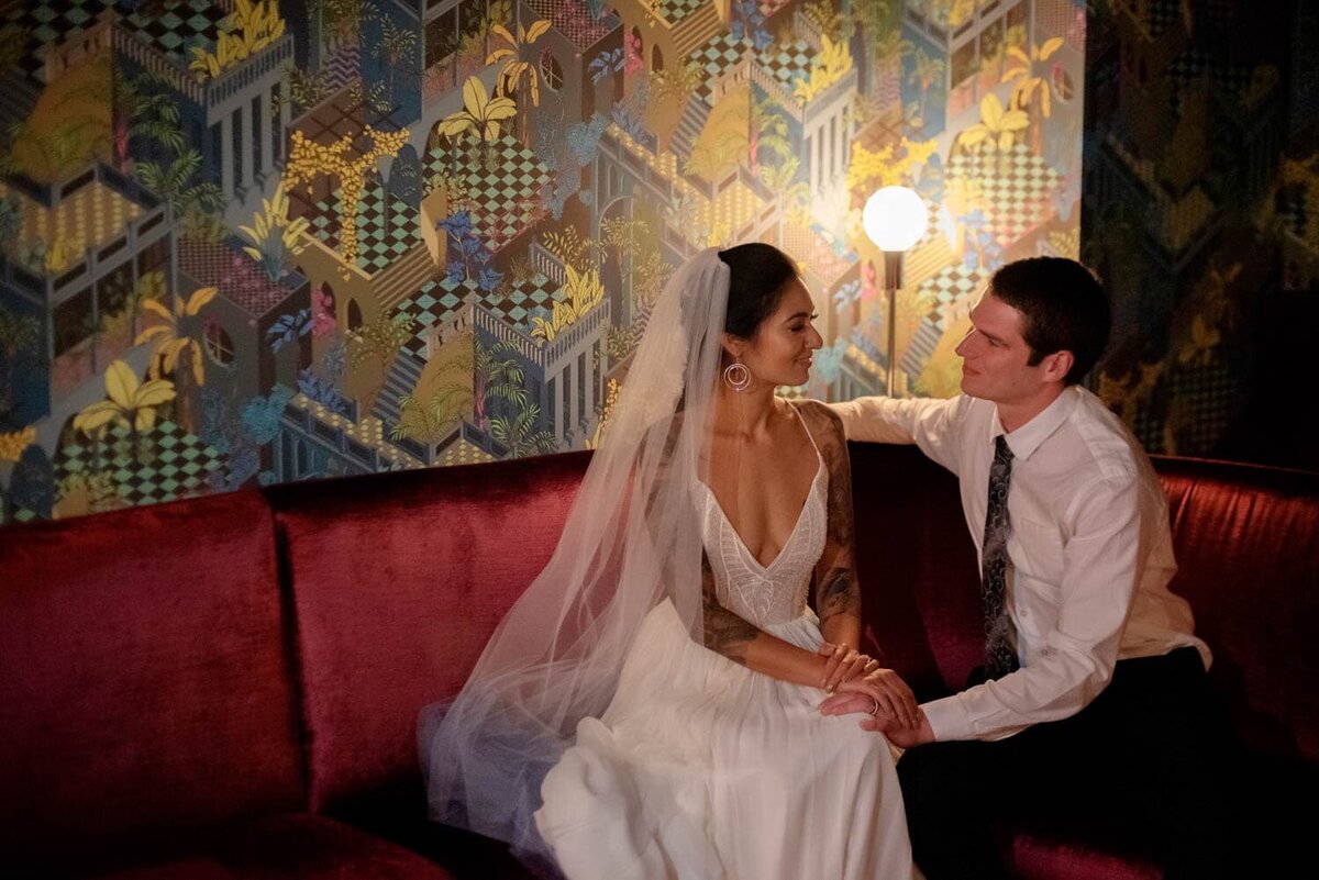 a bride and groom have a moment sitting on a red velvet couch in front of a colorful fun wall in Voysey speakeasy