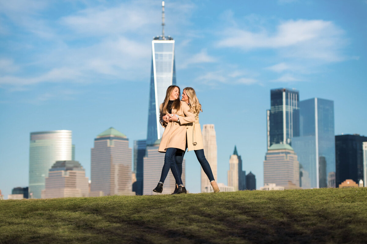 Two women wearing matching jackets, hugging each other in front of the New York City skyline in liberty State Park