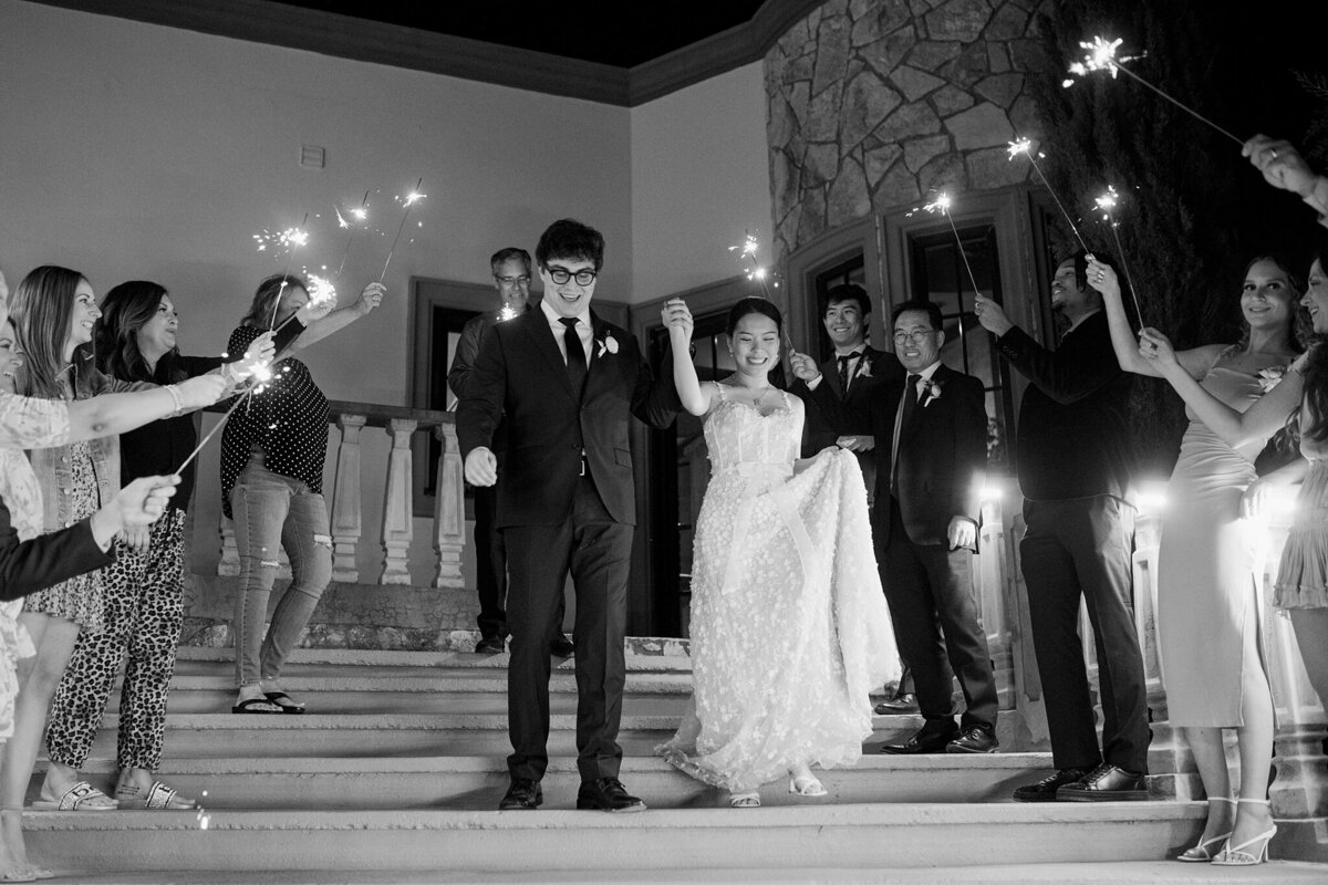 A black and white shot of a bride and groom joyously exiting their wedding reception in DFW, Texas. The bride is on the right and is wearing a long, flowing, detailed, white dress. The groom is on the left and is wearing a dark suit with a boutonniere. Guests form two long lines on either side of them as said guests hold up lit sparklers in celebration.