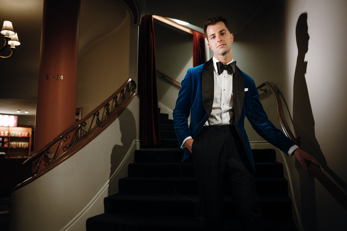 Groom in velvet suit posing at a staircase as a spotlight shines on him.