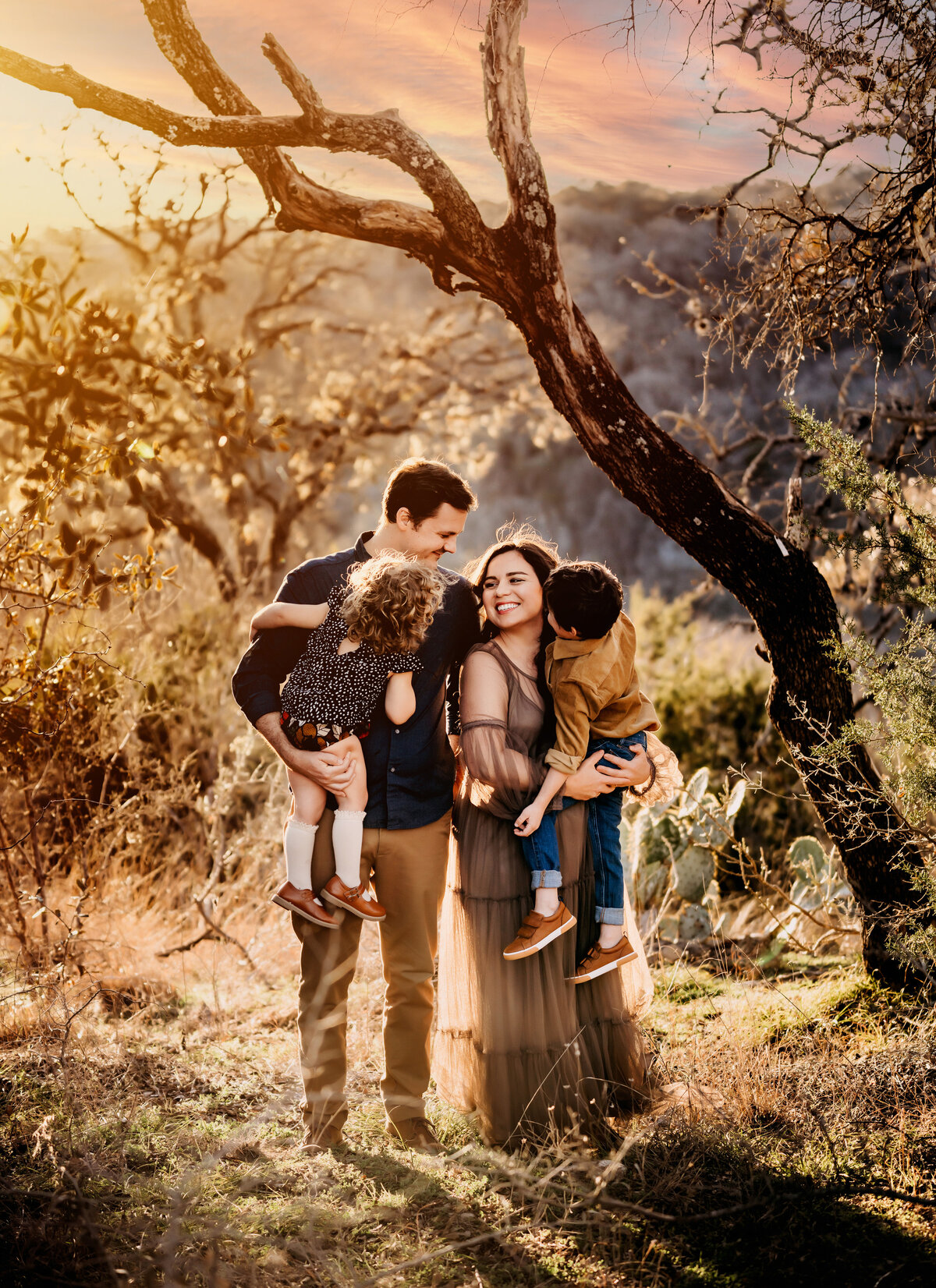 Family Photographer, a young mother and father hold their two young kids beneath tree cover