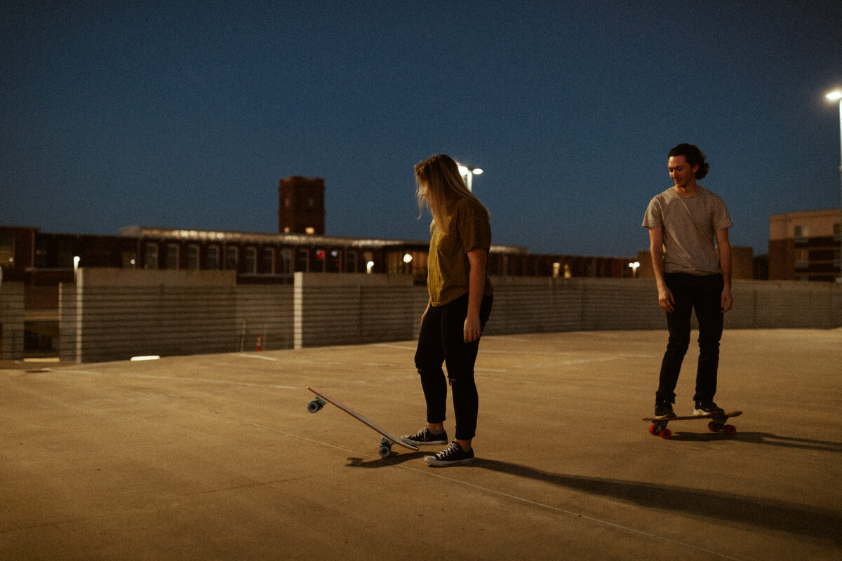A couple is riding their skateboards on top of a parking garage at night under the street lights.