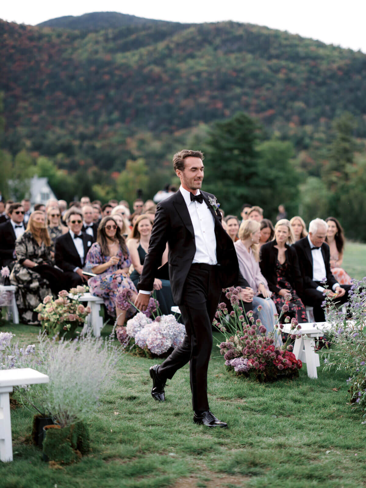 The groom is walking on the wedding aisle outdoors, as the guests watched, at The Ausable Club, NY. Image by Jenny Fu Studio.