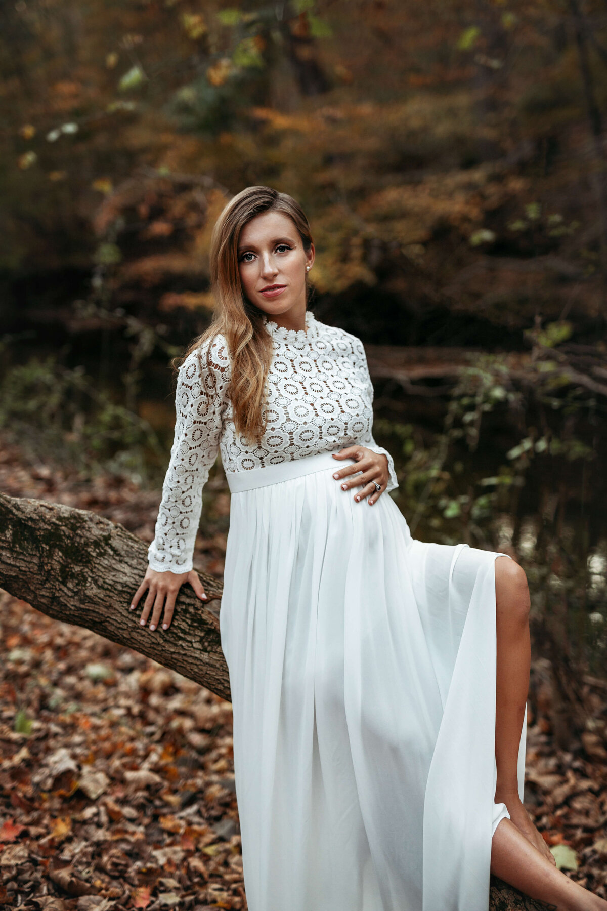 pregnant woman wearing a white maternity gown sitting on a branch in the fall in front of a stream
