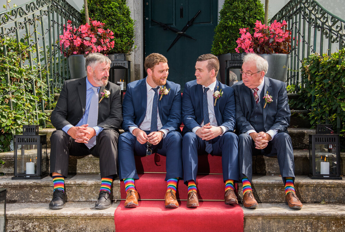 fathers sitting with groomsmen on a step with red carpet and wearing rainbow socks