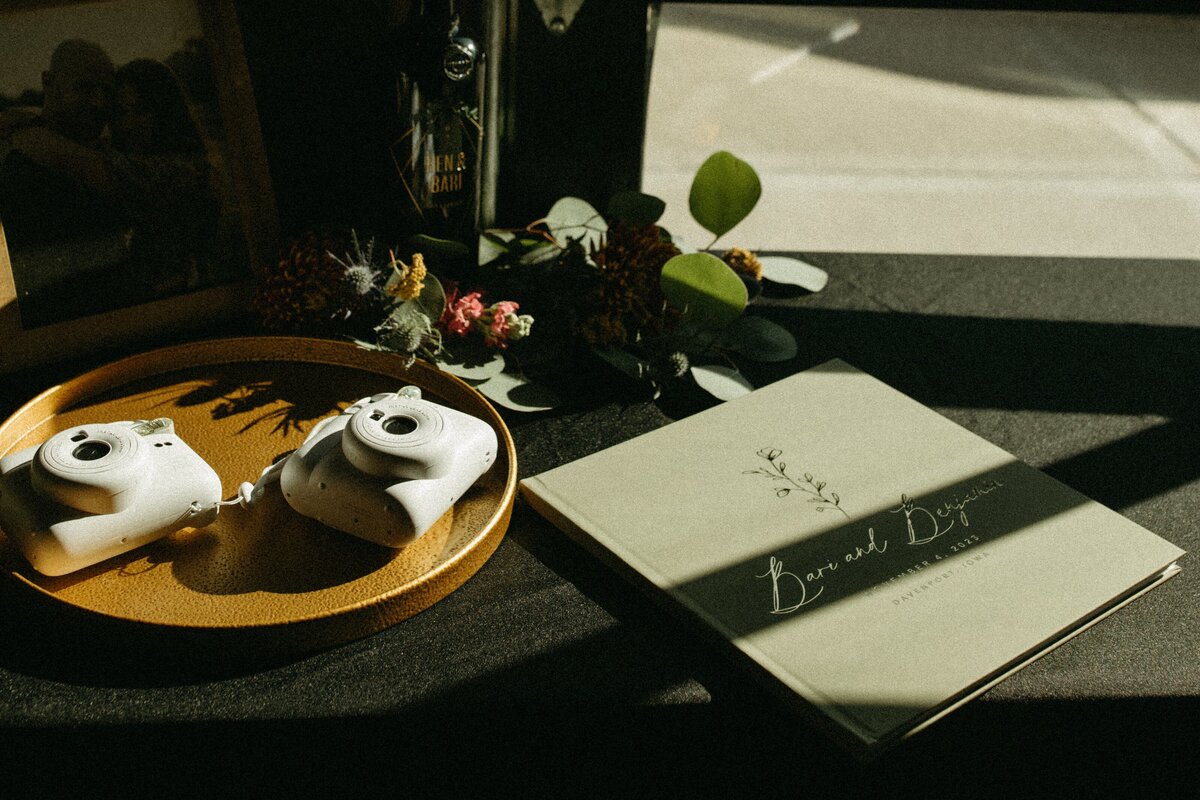 A sunlit table displaying a book titled "Beneath Dirt," two white vintage cameras on a yellow plate, and a small bouquet of dried flowers, perfect for events in Davenport.