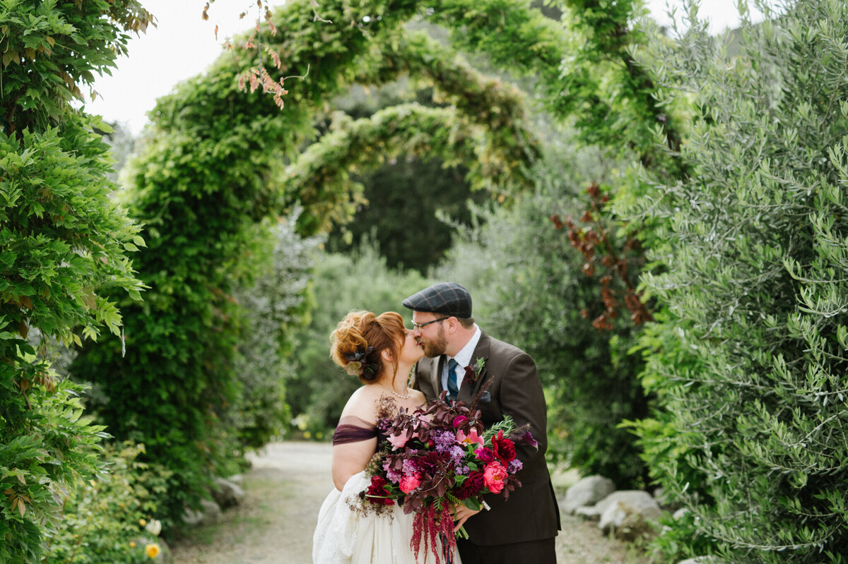 Stylish and unique wedding couple kissing under an arch of trees, gorgeous bride with orange hair holding a stunning large pink and purple bouquet, groom wearing a plaid cap, captured by Christy D. Swanberg Photography, editorial elopement and wedding photographer in Calgary, Alberta, featured on the Bronte Bride Vendor Guide.