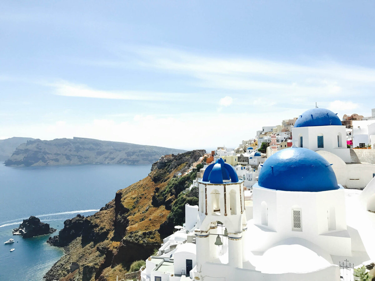 Scenic view of Santorini, Greece, featuring white buildings with blue domes overlooking a calm sea. Steep cliffs extend into the water, with a clear sky enhancing the tranquil atmosphere.