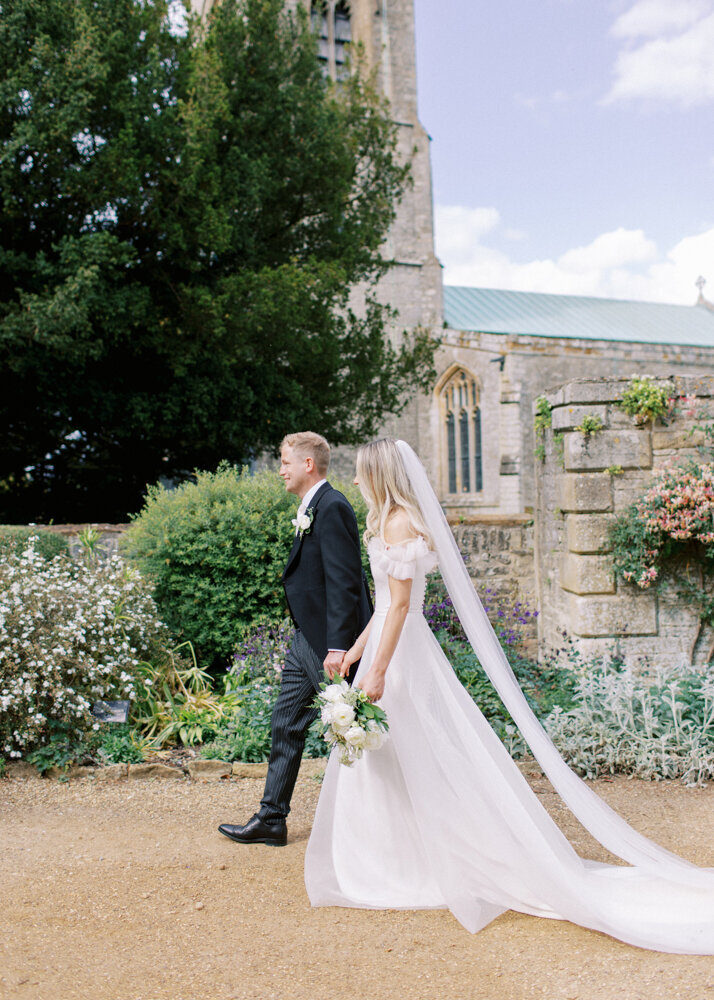 happy bride and groom walking hands to hands in front of St Mary Magdalene church in Castle Ashby
