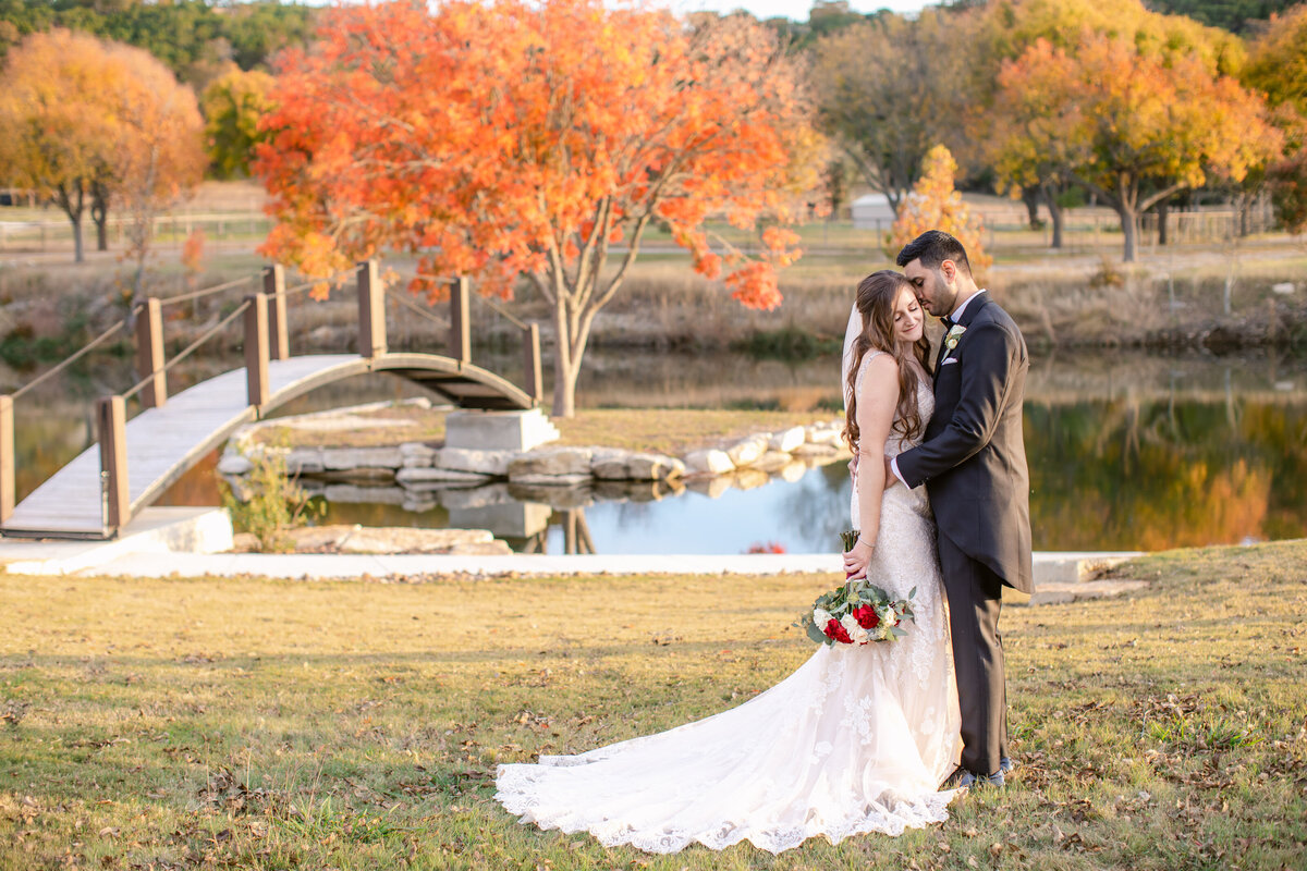 bride and groom embrace with Fall foliage on water's edge behind at Sendera Springs in Kerrville Texas